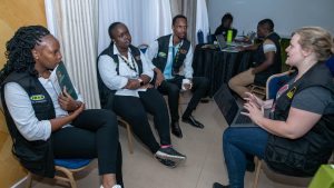 A group of facilitators wearing IRC vests sits in a circle conversing in a conference room.