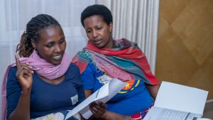 Two women wearing brightly colored scarves around their necks look over work booklets