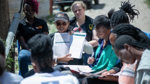 A dozen women sit in chairs arranged in a circle on a dirt road. The facilitator holds up a work booklet while participants listen and follow along in their own booklets.